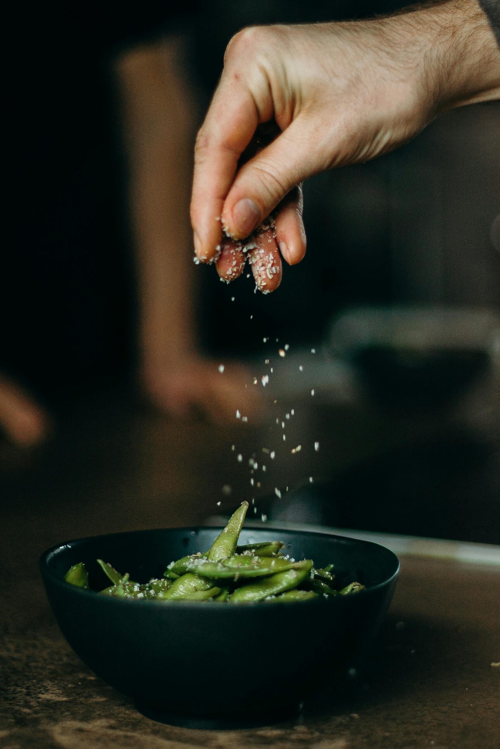 A hand sprinkles salt over a bowl of fresh edamame beans, showcasing a close-up culinary moment.